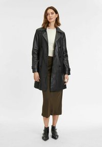 Trench coat in pelle nera con design doppiopetto, rever a disegno e tasche laterali. Il modello indossa un maglione chiaro e una gonna verde oliva.