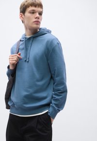 Sweatshirt azul de algodão com capuz ajustável, punhos e bainha canelados, usado com uma mala preta. Camiseta branca visível na parte inferior.