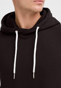 Sudadera negra con una textura suave, que cuenta con una gran capucha y cordones blancos, asegurados con puntas de metal plateado.