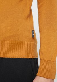 Suéter de punto color mostaza con puños y dobladillo acanalados. La tela es suave y tiene una etiqueta lateral que muestra la marca "ZIGN." Pantalones negros visibles debajo.