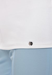 White cotton t-shirt with a curved hem, featuring a small black metallic logo detail near the bottom, paired with light blue fitted pants.