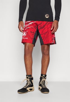 Mann trägt rot-schwarz gemusterte Sportshorts, schwarzen Langarmshirt und schwarz-beige High-Top-Sportschuhe, steht vor hellem Hintergrund.