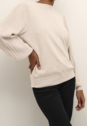 Pullover beige clair avec des manches texturées et côtelées et un corps uni. Il a un col rond et une taille bien ajustée. Associé à un pantalon foncé.