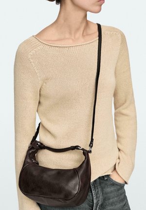 Frau trägt einen beigen Strickpullover und graue Jeans, mit einer dunkelbraunen Leder-Umhängetasche, die von ihrer Schulter hängt.