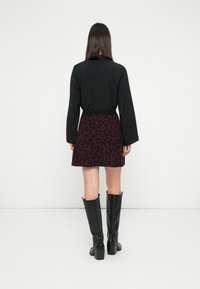 Mujer de espaldas que lleva una blusa negra de manga larga, una falda corta de leopardo morado y botas altas negras con tacón, sobre un fondo blanco.