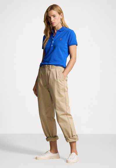 Blue polo shirt with a collar and three-button placket, paired with beige pleated trousers. The model wears white sneakers.
