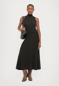 Top negru cu model canelat, fără mâneci, cu un guler înalt, asortat cu o fustă maxi neagră, mulată; model care ține o geantă mică, rotundă, neagră.