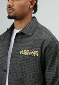Grijs geruit overhemd met een knoopsluiting, grote kraag en gele geborduurde tekst "FREE DOM" aan de linkerkant.