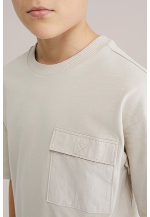 Krupni plan osobe koja nosi svijetlobéž majicu s teksturiranim džepom na prsima i rebrastim okruglim izrezom.