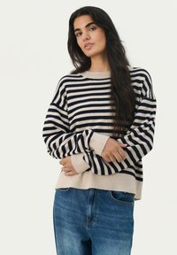 Pull rayé avec des couleurs bleu marine et crème, poignets côtelés et coupe décontractée ; porté avec un jean en denim bleu. Texture en tricot douce.