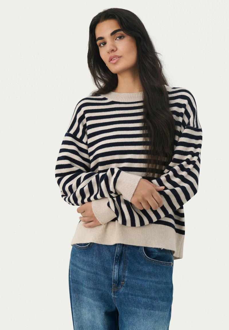 Pull rayé avec des couleurs bleu marine et crème, poignets côtelés et coupe décontractée ; porté avec un jean en denim bleu. Texture en tricot douce.