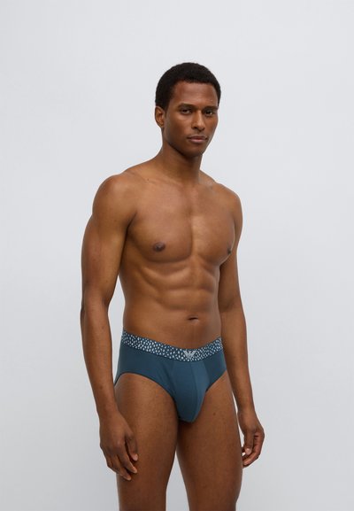 Pánské slipy z tmavě modrozelené látky s páskem s vzorem. Mají hladkou texturu, přiléhavý tvar a pohodlný design.