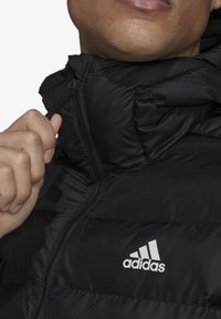 Sort pufferjakke med høj krave, der har lynlåslukning og hvidt Adidas-logo på brystet. Materialet ser skinnende og quiltet ud.