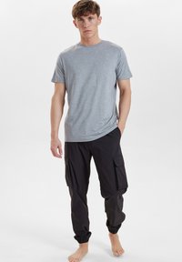 Gray short-sleeve t-shirt, round neck, paired with black cargo pants featuring side pockets and elastic cuffs at the ankles. Barefoot.