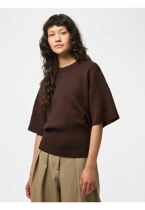 Femme aux longs cheveux bouclés portant un pull ample à manches courtes marron foncé et un pantalon plissé beige, debout devant un fond blanc uni.