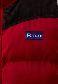 Crvena izolirana jakna s crnim baršunastim akcentima. Ima plavu oznaku s "Penfield" logom koji prikazuje dizajn planinskog obrisa.
