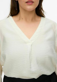 Blouse blanche texturée avec un col en V et un petit détail de bouton à l'avant, associée à une ceinture noire, présentant un tissu doux et fluide.