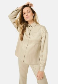 Chemise en satin beige à manches longues, avec un devant à boutons et une poche poitrine unique. Texture lisse, coupe décontractée et détail d'ourlet arrondi.