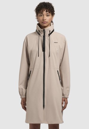 Veste légère beige avec un col montant, cordons de serrage réglables, fermeture éclair à l'avant et deux poches latérales, avec des accents noirs et un logo.