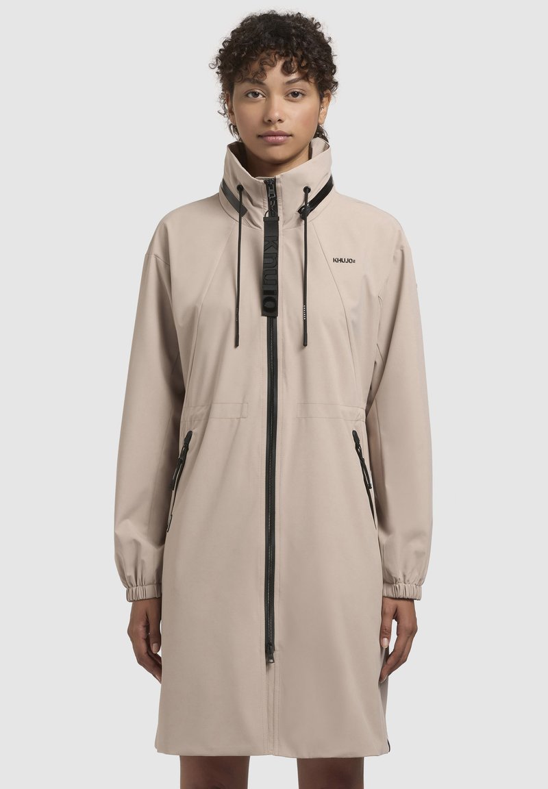 Giacca leggera beige con colletto alto, cordini regolabili, zipper frontale e due tasche laterali, con dettagli neri e branding.