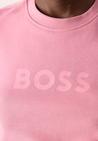 Gros plan d’une personne portant un sweat-shirt rose avec le mot BOSS imprimé en lettres roses un peu plus foncées sur la poitrine.