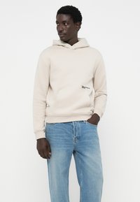 Felpa beige chiaro con una tasca a marsupio e cappuccio con coulisse, dotata di un logo applicato sul lato sinistro, abbinata a jeans azzurri chiari.