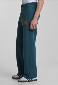 Pantalones de chándal color teal con una cinturilla elástica, bolsillos laterales y un logotipo bordado en naranja en el muslo izquierdo. Combinados con zapatillas grises y blancas.