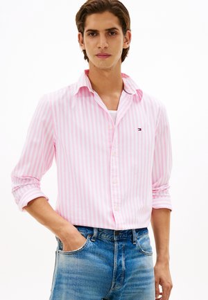Jonge man draagt een roze-wit gestreept overhemd met knopen, opgerolde mouwen en een blauwe spijkerbroek, hand in de zak, tegen een witte achtergrond.