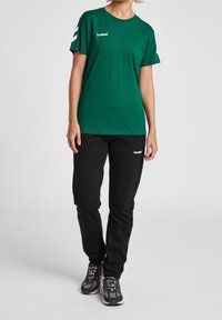 Camiseta de algodón verde con mangas cortas y logo blanco en el pecho, combinada con pantalones deportivos negros y zapatos deportivos grises con detalles.