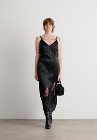 Schwarzes Satin-Nachthemd mit dünnen Trägern, das ein transparentes Spitzenpanel am Rock aufweist. Das Modell hält eine kleine schwarze Handtasche und trägt kniehohe Stiefel.