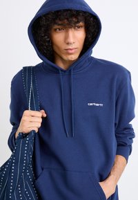 Navy blue hoodie features a kangaroo pocket, drawstring hood, and white "Carhartt" logo on the chest. The model holds a patterned bag.