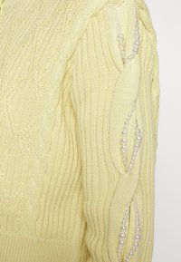 Maglione giallo a maglia con texture a coste, caratterizzato da perle decorative lungo le cuciture laterali e un classico scollo rotondo.