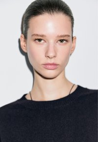 Primo piano del volto di una persona con pelle chiara, occhi nocciola e capelli tirati indietro. Indossa un maglione scuro con scollatura rotonda.