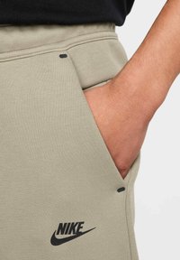 Helle olivfarbene Sweatpants aus strukturiertem Stoff, mit seitlicher Tasche und einem schwarzen Nike-Logo in der Nähe der Tasche. Schwarze Akzente.