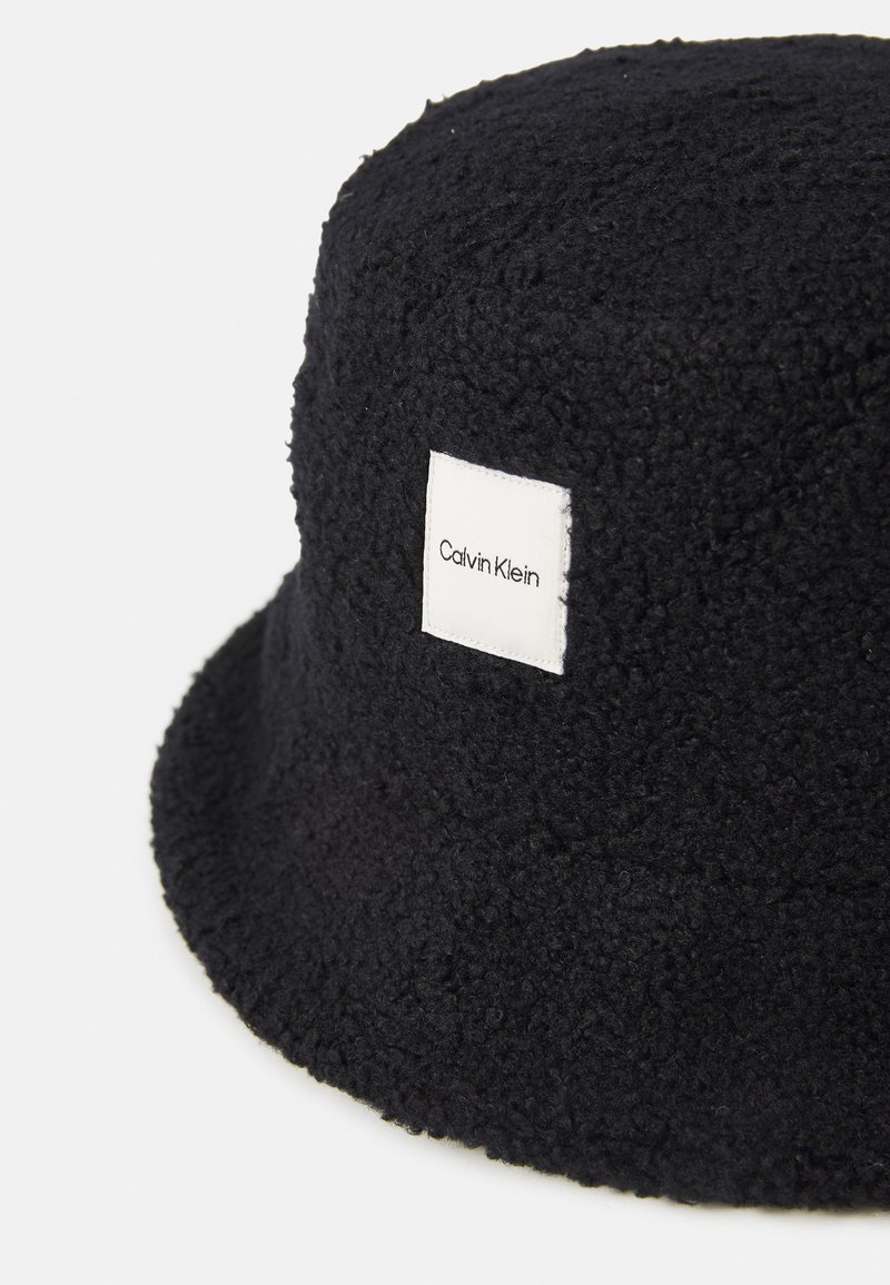 Chapeau seau noir texturé avec un patch logo Calvin Klein blanc centré à l'avant, affiché sur un fond blanc uni.