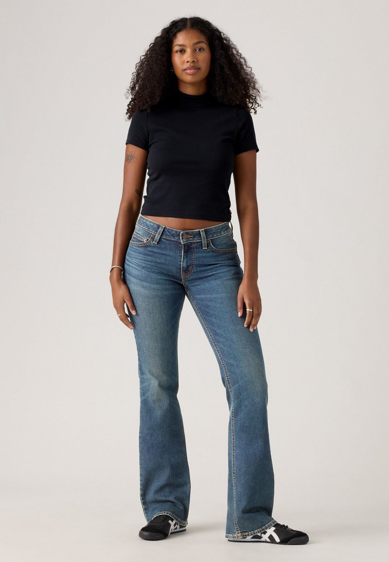 Woman with curly hair wearing a black fitted short-sleeve top, blue flared jeans, black sneakers, and standing against a neutral background.