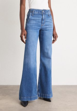 Personne portant un jean évasé bleu clair taille haute avec un top blanc et des chaussures noires à bout pointu, debout sur un sol beige.