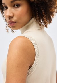 Νεαρή γυναίκα με σγουρά μαλλιά που φοράει μπεζ αμάνικη μπλούζα με ψηλό γιακά, κοιτάζοντας πάνω από τον ώμο της σε απλό φόντο.