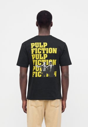 Personne portant un T-shirt noir avec le texte jaune répété "PULP FICTION" et deux hommes en costume tenant des armes dans le dos, pantalon beige visible.