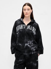 Mulher a usar um hoodie com fecho de correr em tie-dye preto e cinza com o texto "NIGHTMARE" e calças correspondentes, de pé contra um fundo claro e liso.