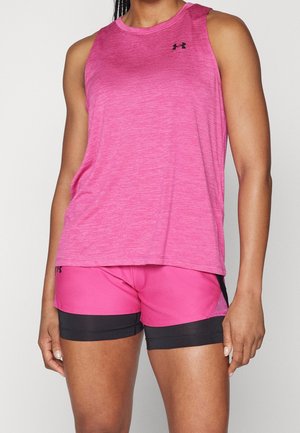 Vrouw draagt een roze mouwloos sporttopje en roze-zwarte sportshorts, staand tegenover een lichte achtergrond.