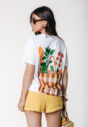 Femme portant des lunettes de soleil et une chemise blanche avec une illustration de sac de course et le texte « Colourful Rebel », associée à un short en tricot jaune.