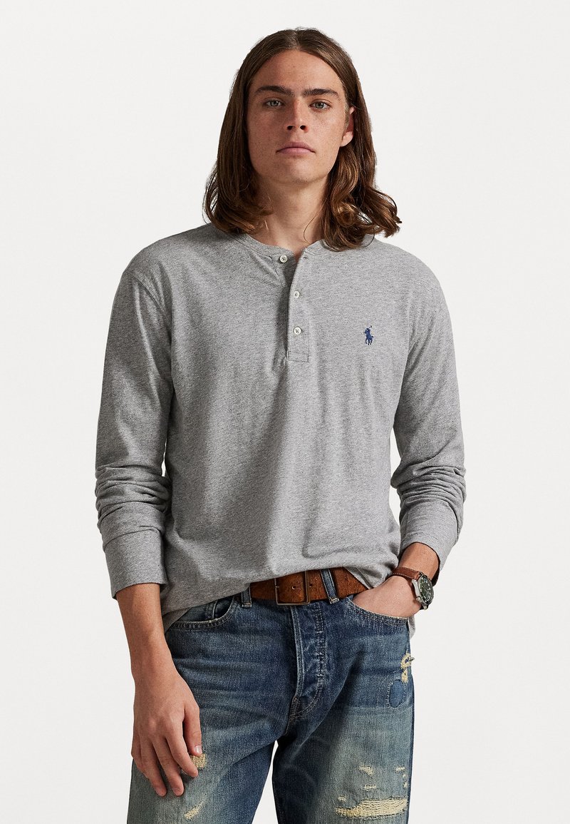 Γκρι μακρυμάνικη μπλούζα τύπου henley με τρία κουμπ�ιά, με μικρό μπλε λογότυπο στο αριστερό στήθος, συνδυασμένη με σκισμένα μπλε τζιν και καφέ ζώνη.