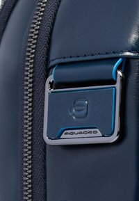 Bolso de cuero azul marino con cremallera plateada, que presenta una etiqueta metálica con detalles azules y el logo "Piquadro". Textura suave, diseño elegante.