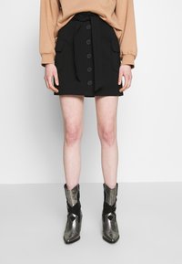 Jupe boutonnée noire avec taille nouée, dotée de poches avant. Portée avec des bottines métalliques à imprimé serpent et bouts pointus.