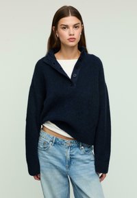 Navy blue knitted pullover sweater with a high collar, three buttons, and a relaxed fit. Paired with light blue denim jeans.
