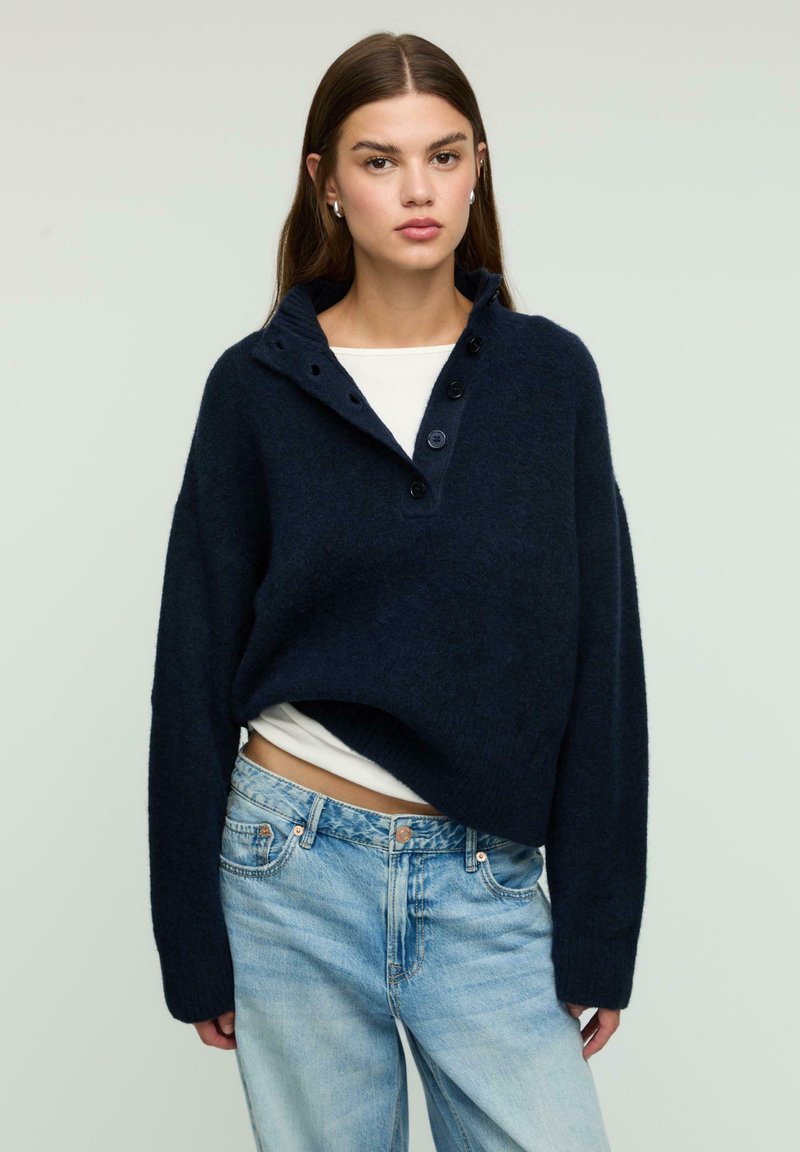Navy blue knitted pullover sweater with a high collar, three buttons, and a relaxed fit. Paired with light blue denim jeans.
