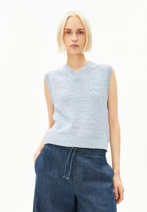 Jeune personne aux cheveux blonds courts portant un pull sans manches tricoté bleu clair et un pantalon ample bleu foncé, les mains dans les poches.