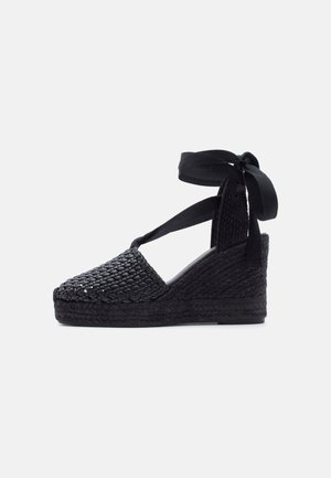Sandală cu platformă neagră, cu textură țesută, vârf închis și baretă cu legătură în jurul gleznei, pe un fundal deschis.