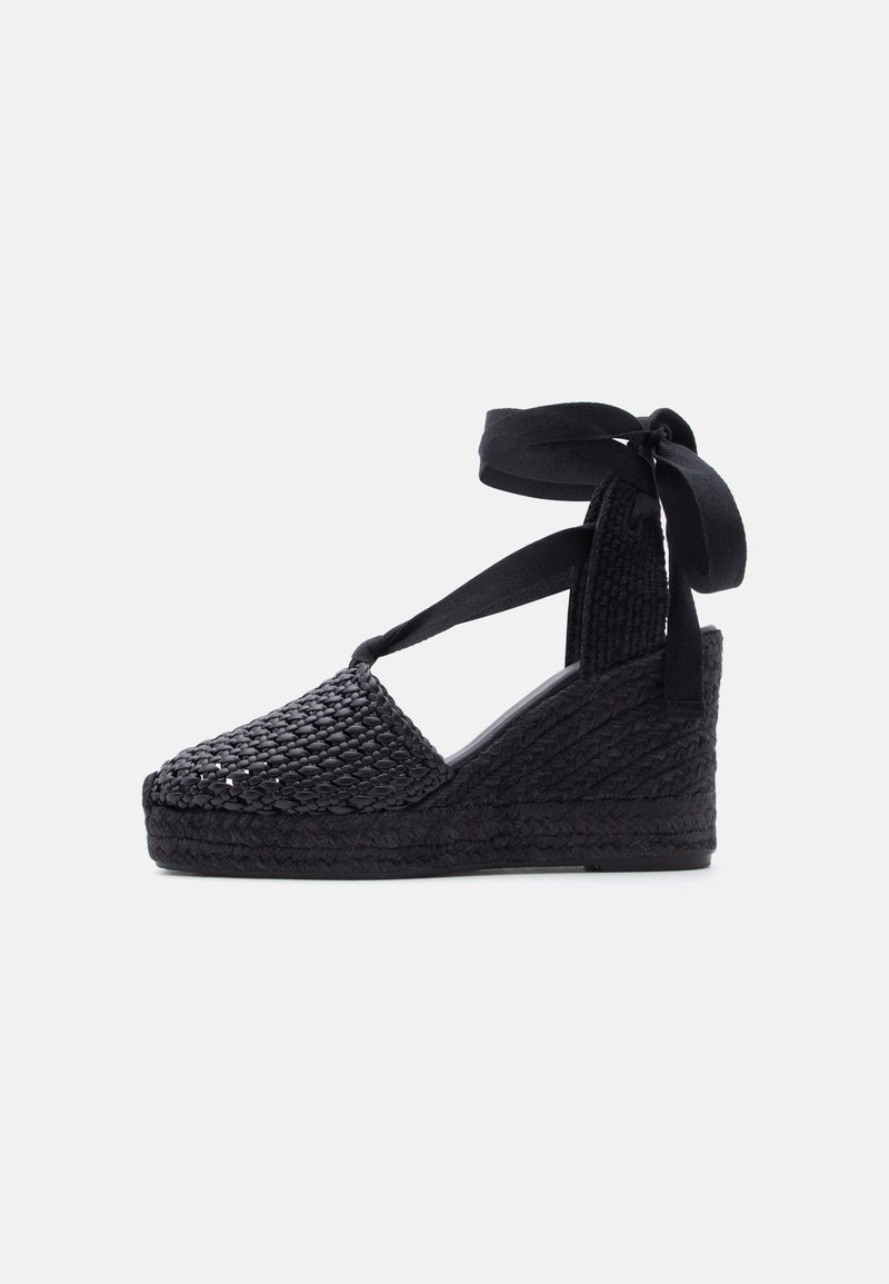 Sandale à semelle compensée noire avec texture tissée, bout fermé et bride nouée à la cheville sur fond clair.